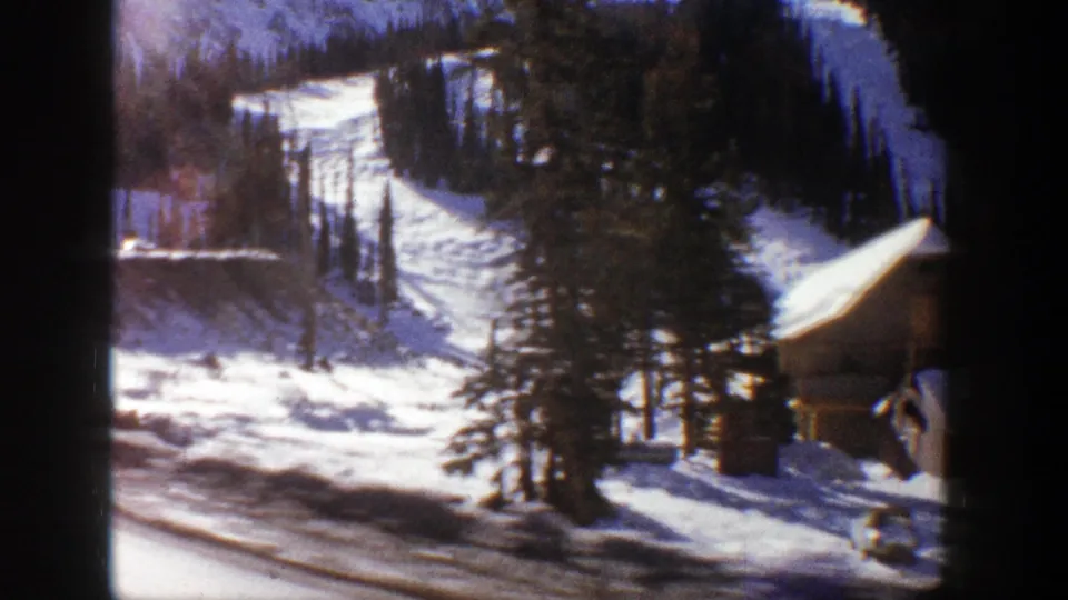 Aspen-area winter slope on 8mm film, showing snow, dense trees, road edges, and a small building partly visible.