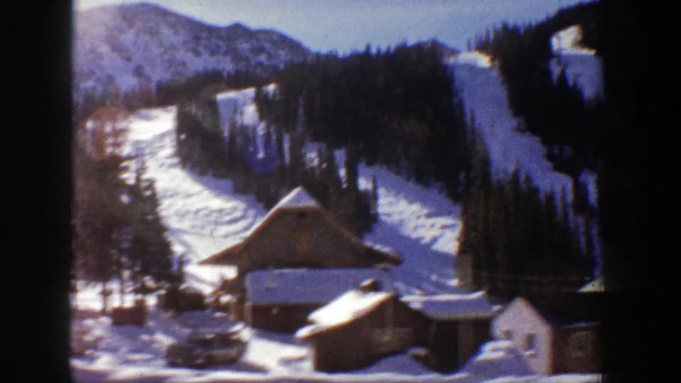 Aspen winter scene from 1961 with cabins, parked cars, evergreen trees and bright snow on a mountain slope.