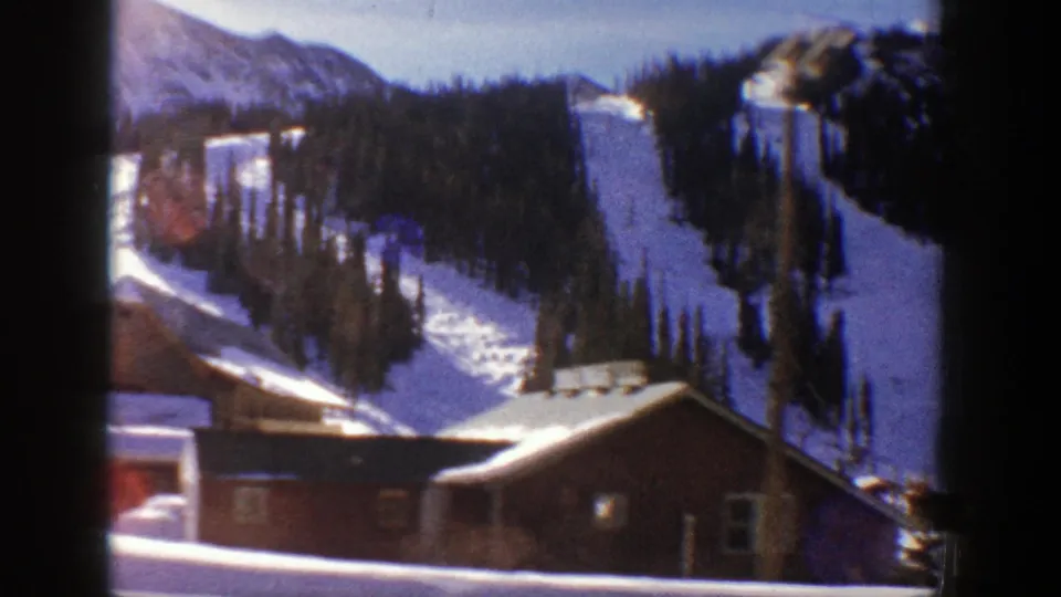 1961 Aspen 8mm frame with a cabin roof in the foreground, dark evergreens and steep snow-covered mountain terrain behind.