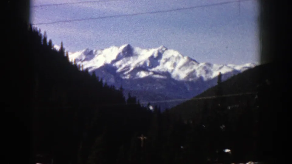 1961 Aspen, Colorado home-movie frame looking toward snow-covered peaks beyond dark evergreen hills, with visible 8mm frame borders.