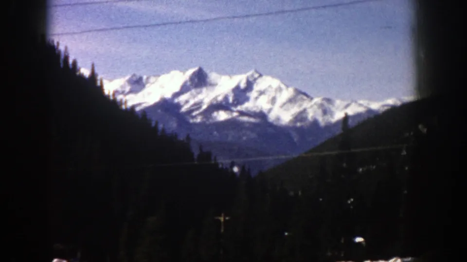 1961 Aspen, Colorado view of distant snow-bright peaks behind dark evergreen slopes, with soft home-movie focus and frame edges.