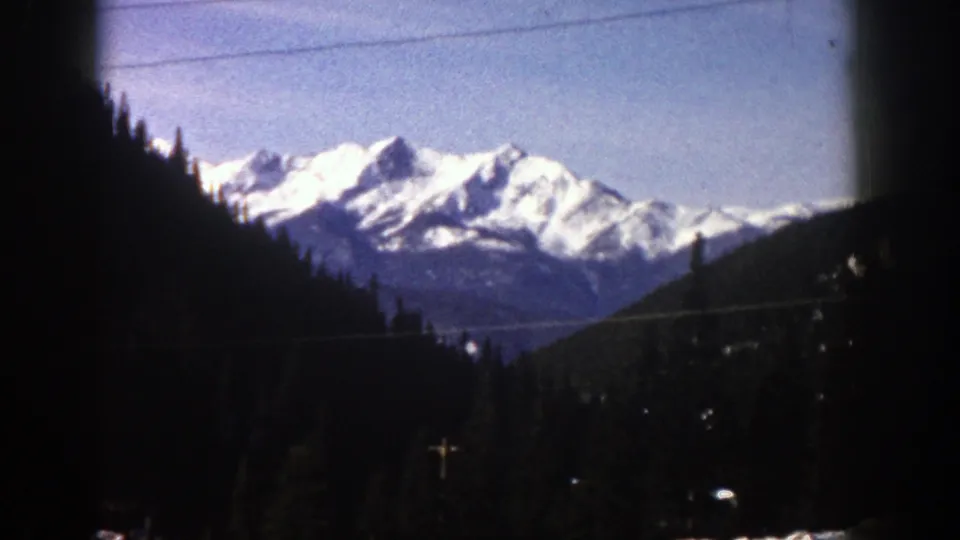 1961 8mm frame from the Aspen, Colorado metadata showing snowy peaks beyond forested slopes, with utility lines across the upper frame.