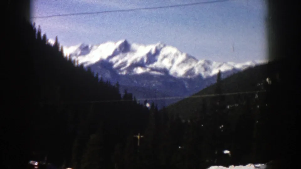 1961 home-movie frame showing snow-capped mountains beyond evergreen slopes, with utility wires and roadside poles visible in the foreground.