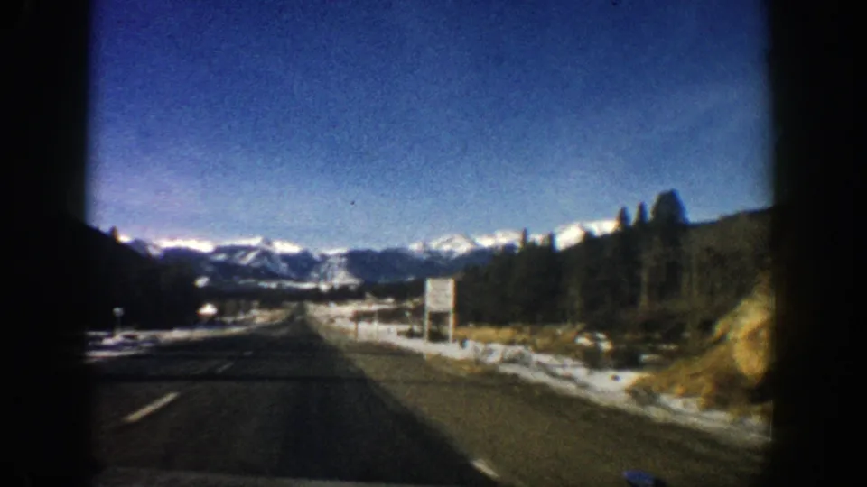Moving-car 8mm view of a two-lane mountain road near the Aspen, Colorado metadata location, with snow patches and distant peaks ahead.