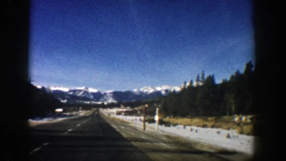 Forward-facing 1961 Aspen-area road view with snow-lined shoulders, evergreen trees, and mountains ahead.