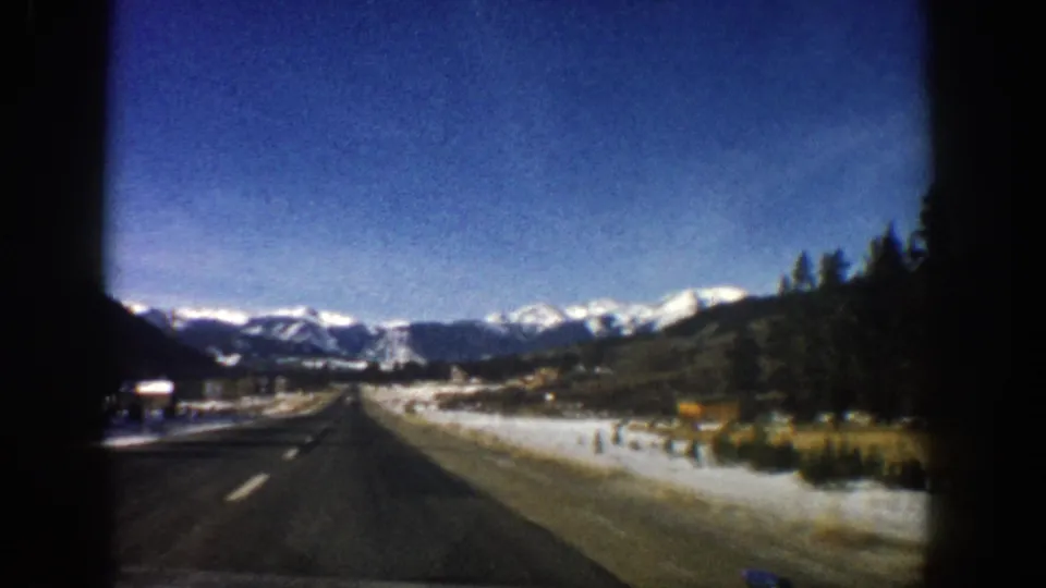 Late-frame 1961 Aspen road view with snow, low roadside signs, evergreens, and distant mountain peaks.
