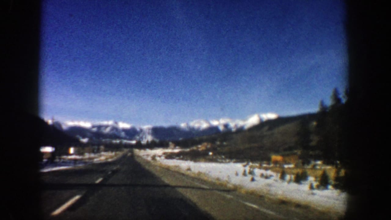 Roadside view through a vehicle window with mountains, snow patches, and evergreen trees ahead.