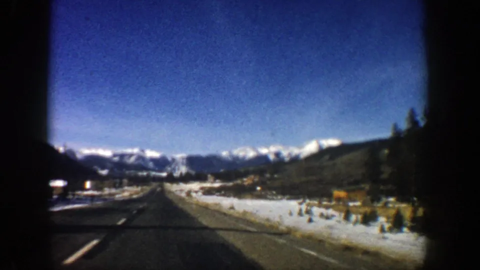 1961 Aspen-area road view through a vehicle window, showing a paved road, snow patches, evergreens, and distant mountains.