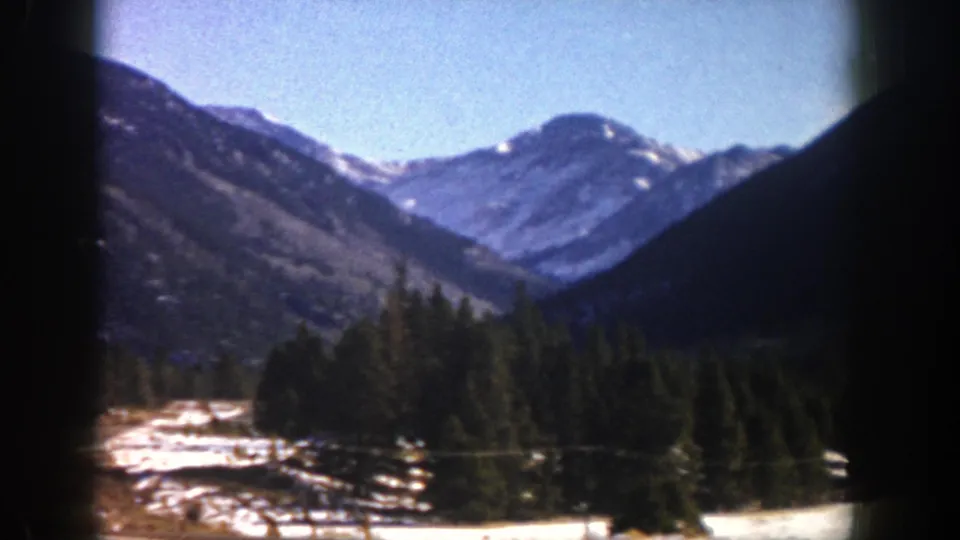 1961 Aspen-area mountain view framed by a vehicle window, with evergreen forest, snow patches, and ridgelines under blue sky.