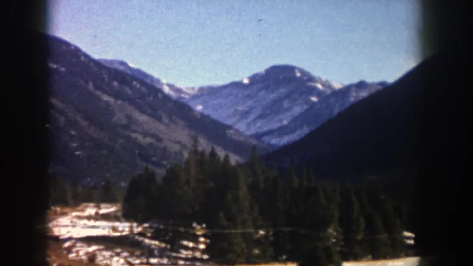 1961 Aspen-area home-movie frame showing a mountain valley, evergreen trees, snow-covered ground, and dark vehicle-window framing.