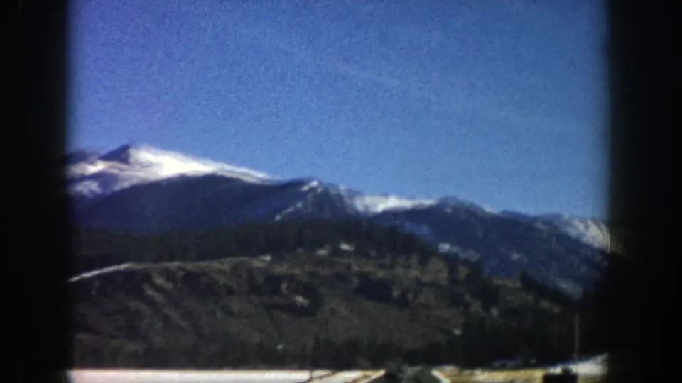 1961 Aspen, Colorado home-movie frame with a snow-covered ridge, open blue sky, dark valley slope, and road at the lower edge.