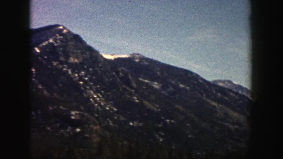 1961 Aspen, Colorado 8mm frame showing a closer dark mountain slope with scattered snow patches beneath blue sky.