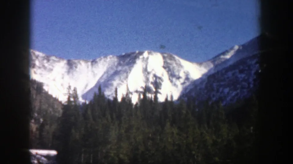 1961 Aspen, Colorado frame showing bright snow-covered mountain peaks above a dense evergreen forest under clear blue sky.