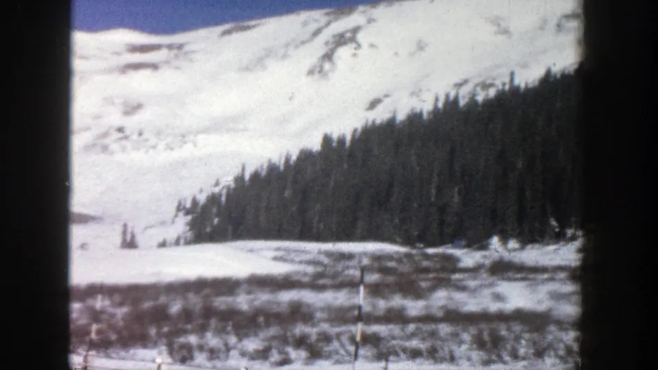 Aspen, Colorado 1961 8mm frame of a snowy mountain slope, evergreen trees, and winter valley foreground seen from a road trip perspective.
