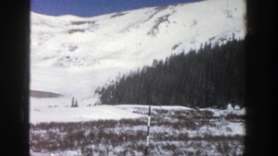 1961 Aspen, Colorado 8mm frame showing a snow-covered mountain, blue sky, evergreen trees, and open winter ground.