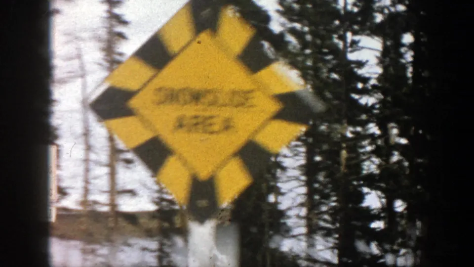 Close roadside view of a yellow warning sign against snowy Aspen trees, with text mostly unreadable in the 8mm frame.
