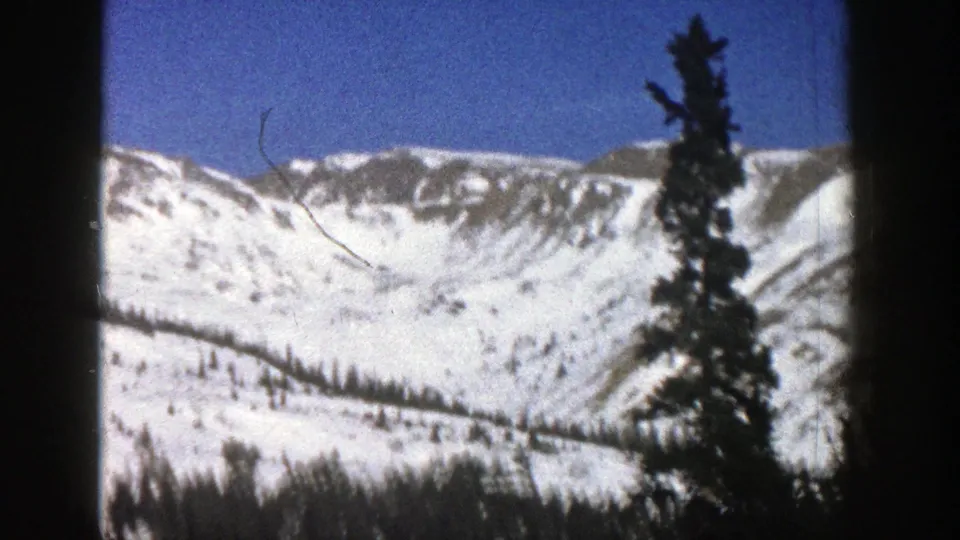 1961 Colorado home-movie frame with snow-streaked peaks, a tall evergreen, blue sky, and visible 8mm frame edges.