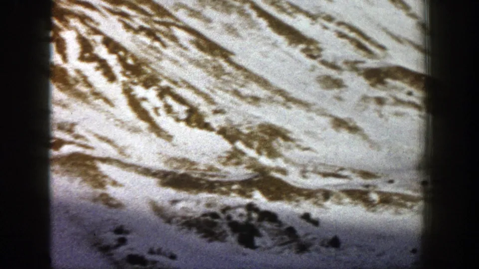 1961 Aspen, Colorado 8mm frame of a rugged snowy mountain face with brown ridges and dark evergreen clusters.