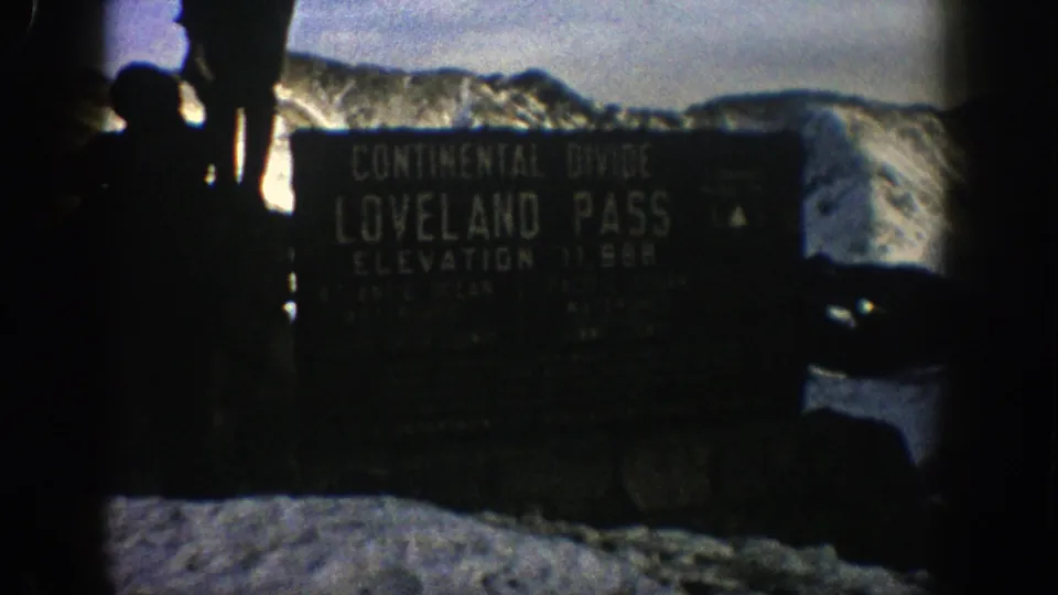 The Loveland Pass Continental Divide marker is framed with dark silhouetted visitors and snow-covered mountains behind it.