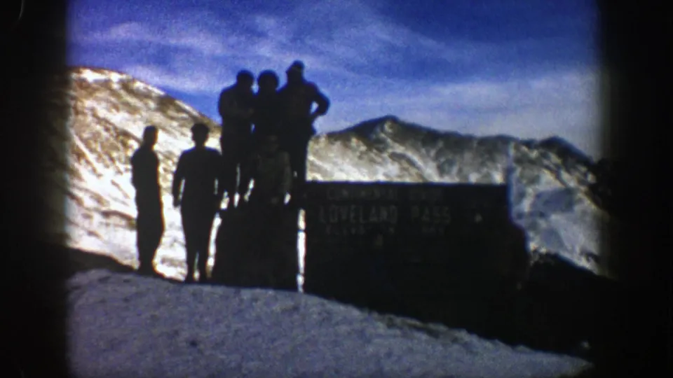 1961 Colorado home-movie frame showing visitors silhouetted on snow beside the Loveland Pass sign and distant mountain peaks.