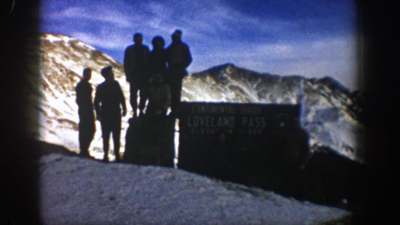 1961 Colorado home-movie frame showing men silhouetted beside the Loveland Pass sign against snow-covered mountain ridges.