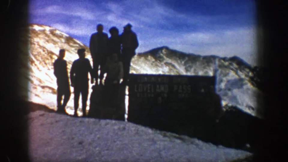 Men stand around the Loveland Pass sign in a backlit 1961 Colorado mountain scene with snow and blue sky.