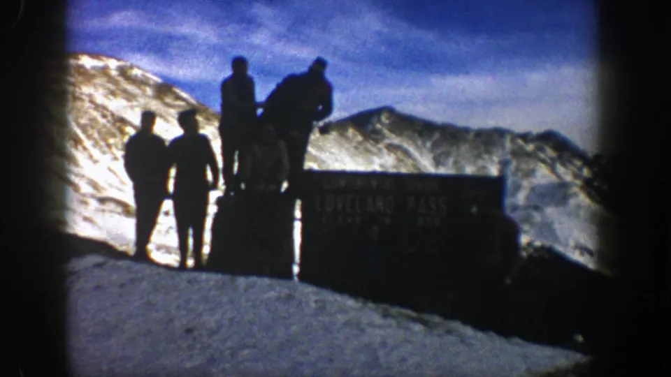 Backlit 1961 Loveland Pass frame with two men in the foreground and others near the sign above snowy mountain scenery.