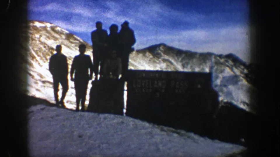 Final 1961 Colorado frame of men silhouetted at Loveland Pass, with the sign partly readable and snowy peaks behind.
