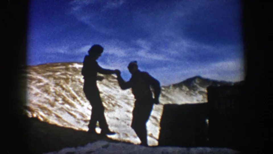 In metadata-supported 1961 Colorado footage, two bundled hikers appear in silhouette while holding hands on a snowy ridge.