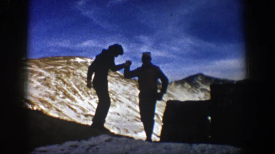 Two hikers in winter clothing stand near a dark sign on a snowy Colorado mountain pass, with ridges and blue sky behind them.