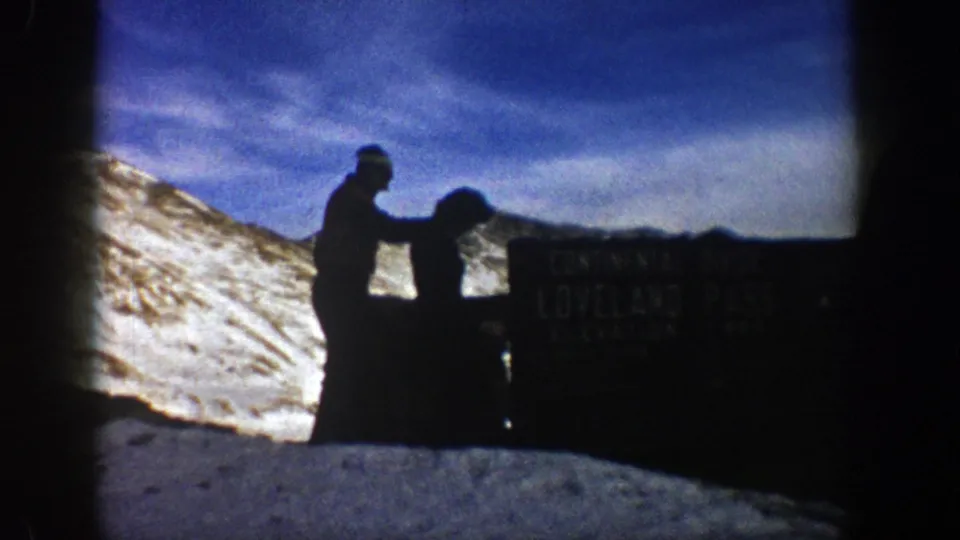 A silhouetted hiker reaches toward a companion beside a partly readable elevation sign in snowy Colorado high country.