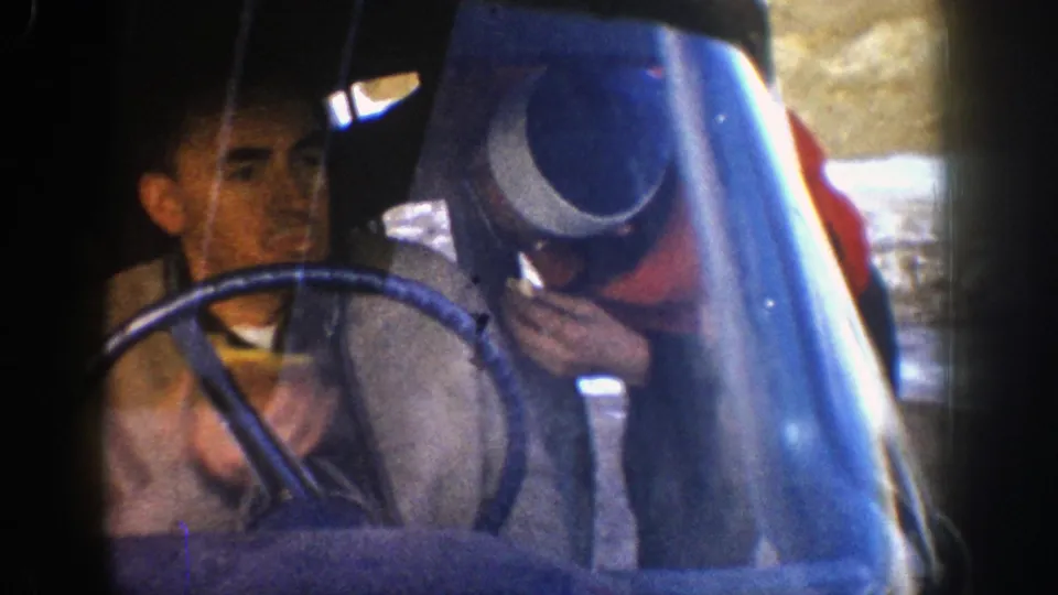 A man in outdoor clothing sits near a steering wheel or vehicle-like structure during the same 1961 Colorado mountain outing.