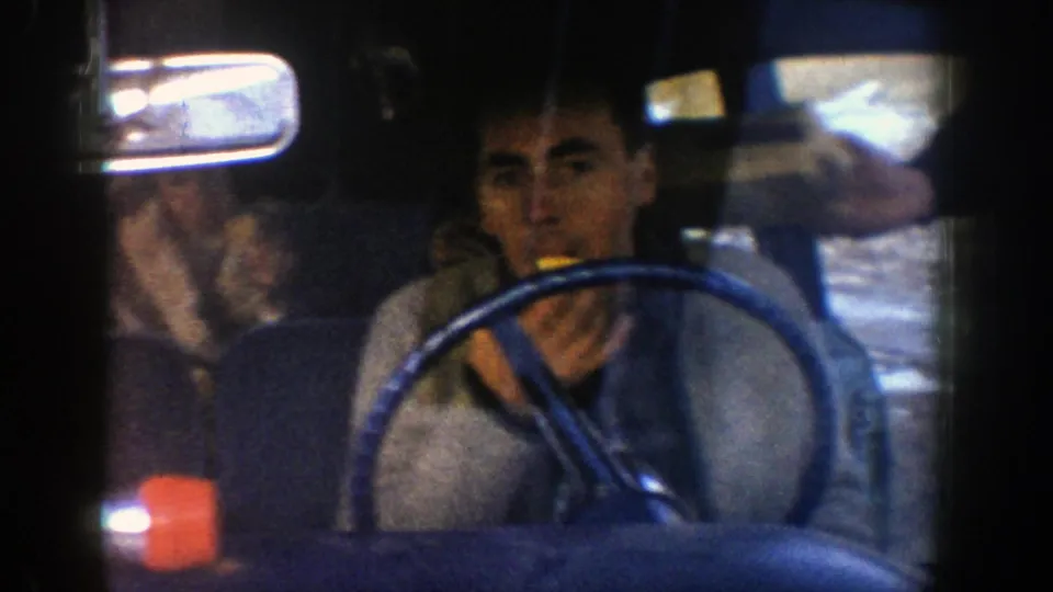 A man sits behind the blue steering wheel of a car in the 1961 Aspen, Colorado footage, raising a small yellow food item while daylight reflects through the windshield.