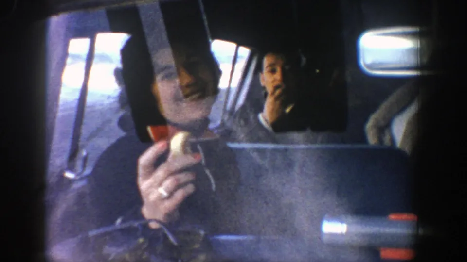 Inside the car, an adult in the front passenger area and another passenger in the rear are visible eating or holding small yellow food items in the 1961 Aspen, Colorado home-movie footage.