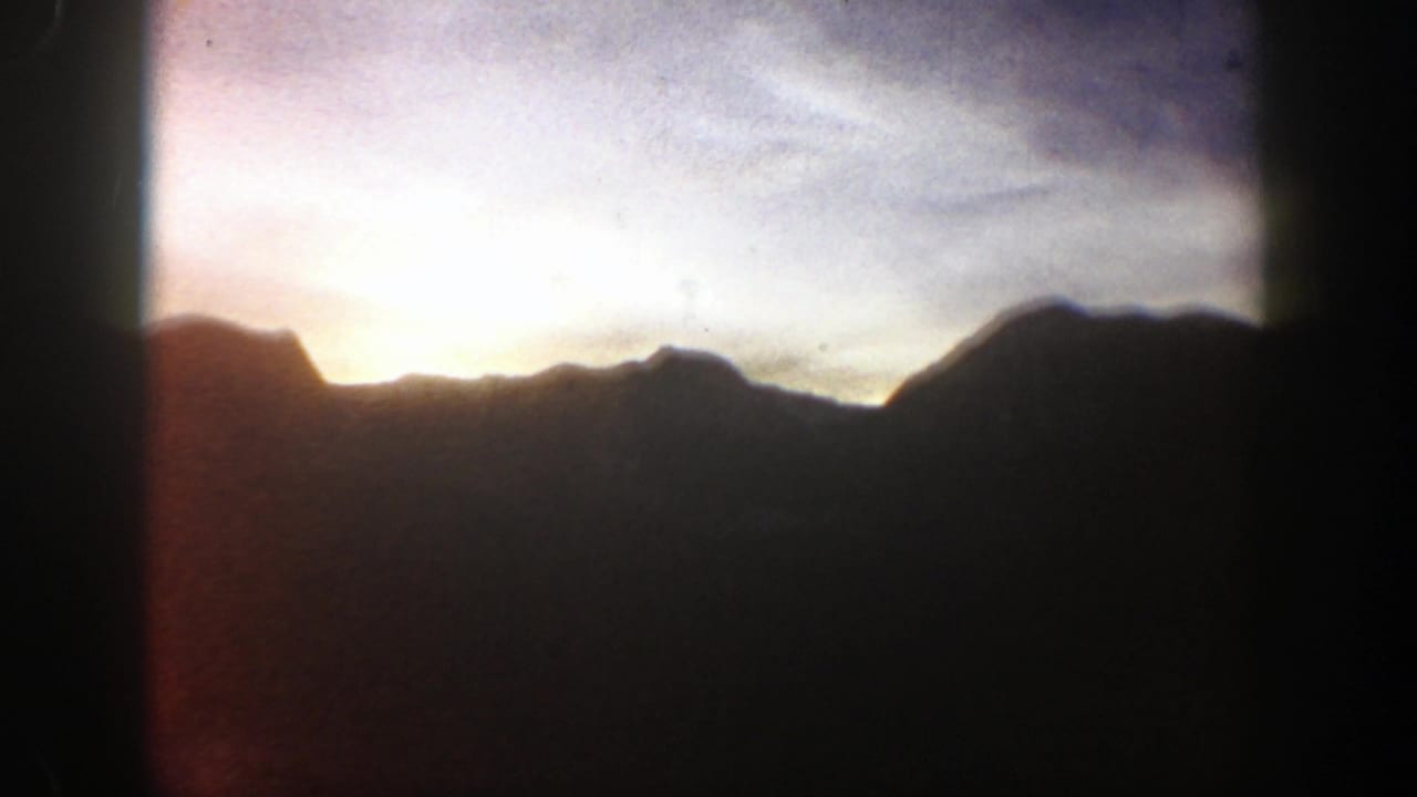 1961 Aspen, Colorado home-movie frame showing a dark mountain silhouette beneath pale dawn clouds and a low sunrise glow.