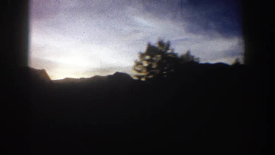 1961 Aspen, Colorado frame with silhouetted mountains, a pale sunrise band, streaked clouds, and a dark tree shape near the ridge.