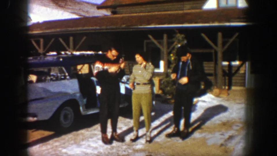 In 1961 Aspen, adults talk near an open white station wagon with roof gear outside a rustic lodge on snowy ground.