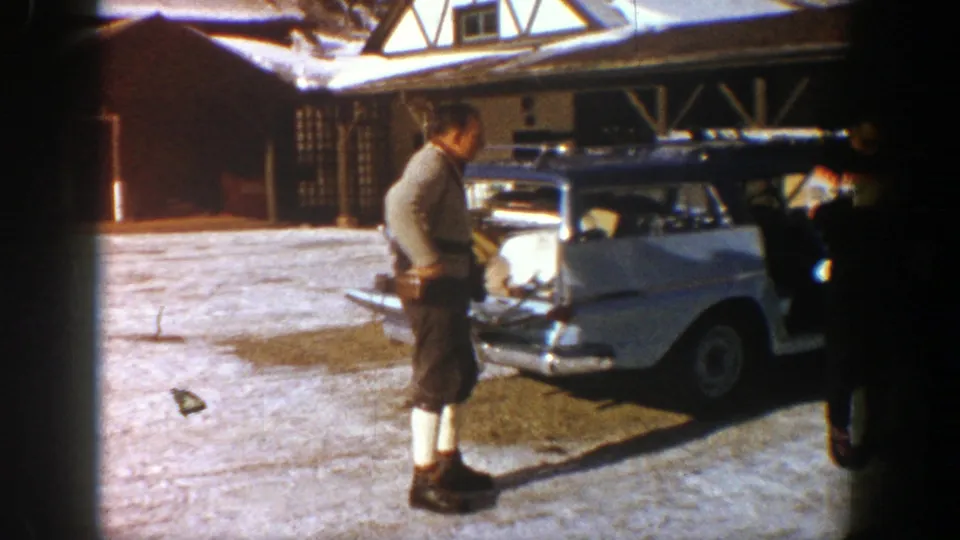 In snowy Aspen, a man wearing alpine-style clothing stands by the rear of a loaded white station wagon near a lodge.