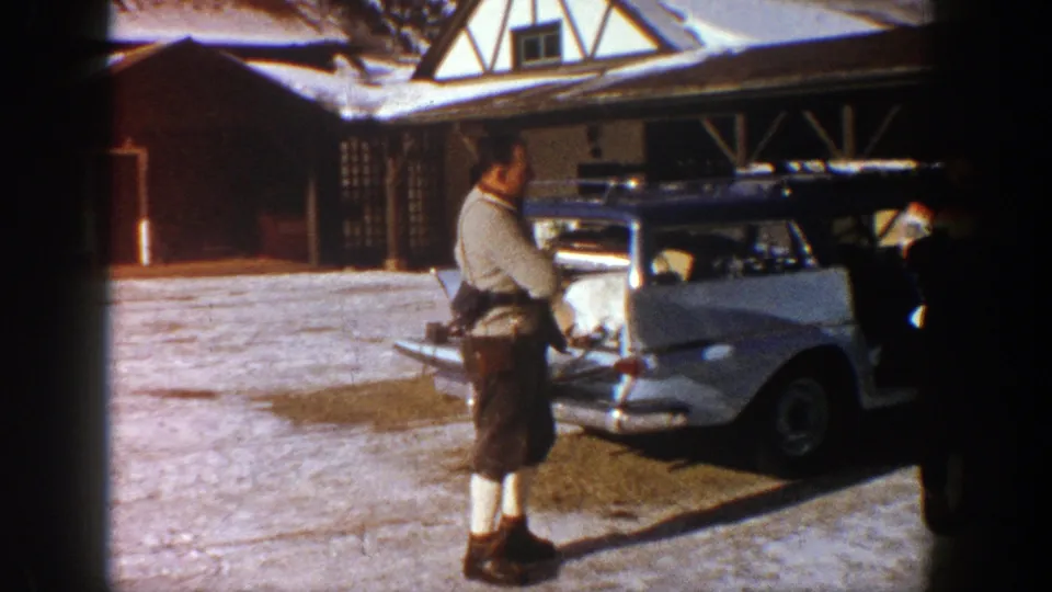 A man in alpine-style dress adjusts his clothing beside a white station wagon outside a rustic Aspen lodge in 1961.