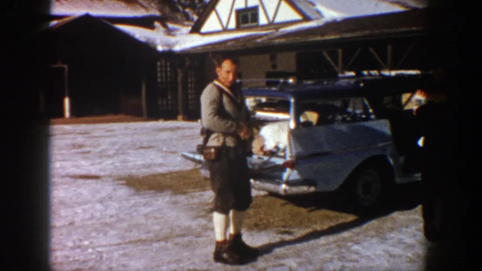 In a 1961 Aspen home movie frame, a man faces the camera near a loaded station wagon outside a rustic lodge.