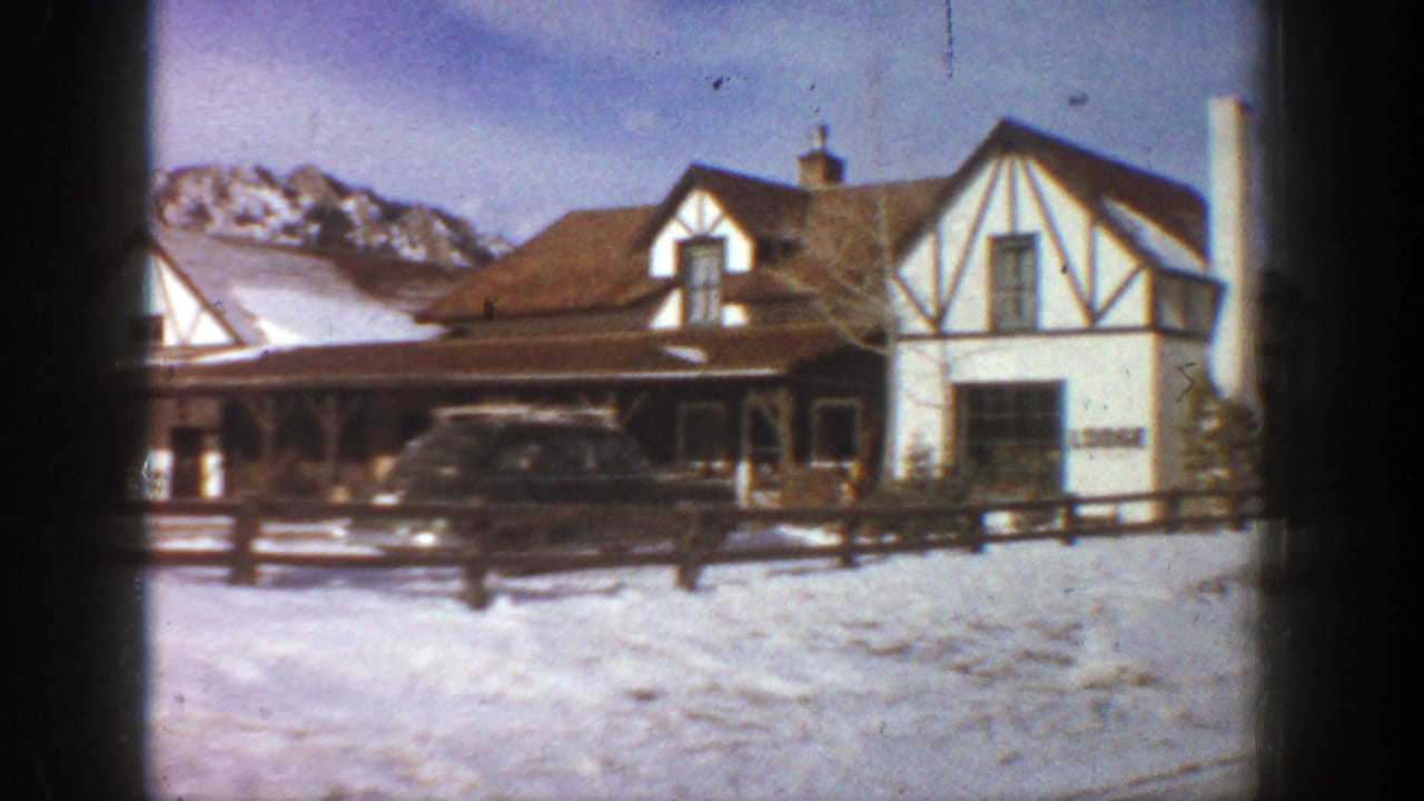 Snowy Tudor-style lodge with a parked station wagon and wooden fence in front.
