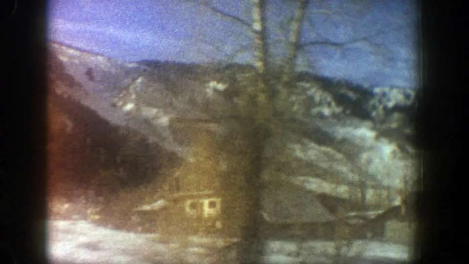 Home-movie frame from Aspen showing cabins, bare trees, snow patches, and mountain slopes in soft 8mm color.