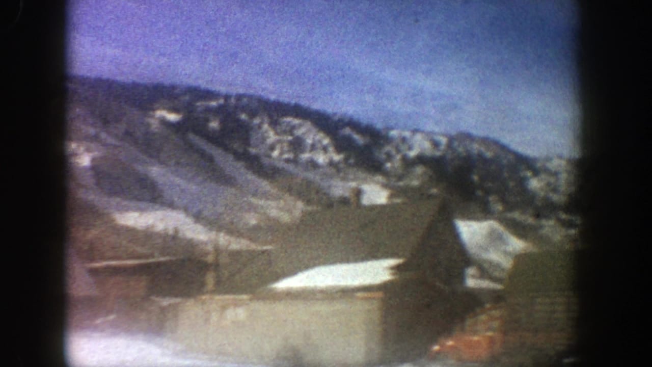 Snowy mountains behind low wooden buildings and a dark film-frame border.
