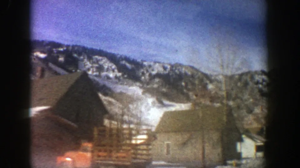 Aspen, Colorado, 1961 frame with wooden structures, a red vehicle, bare branches, and snow-covered mountain terrain.