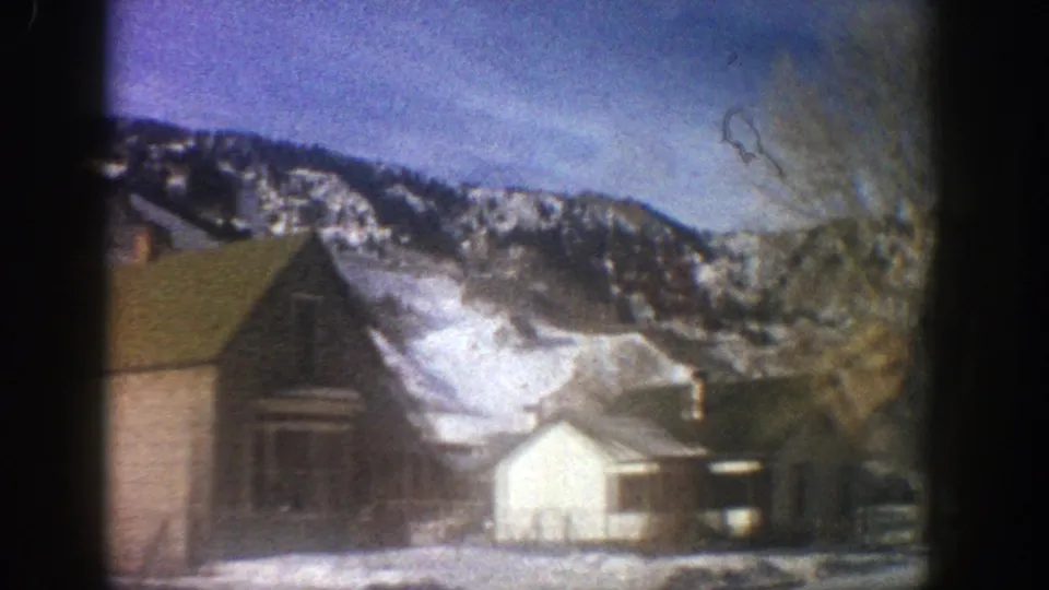 Aspen, Colorado, 1961 frame showing a wooden house, white outbuilding, bare tree, and snow-covered mountain backdrop.