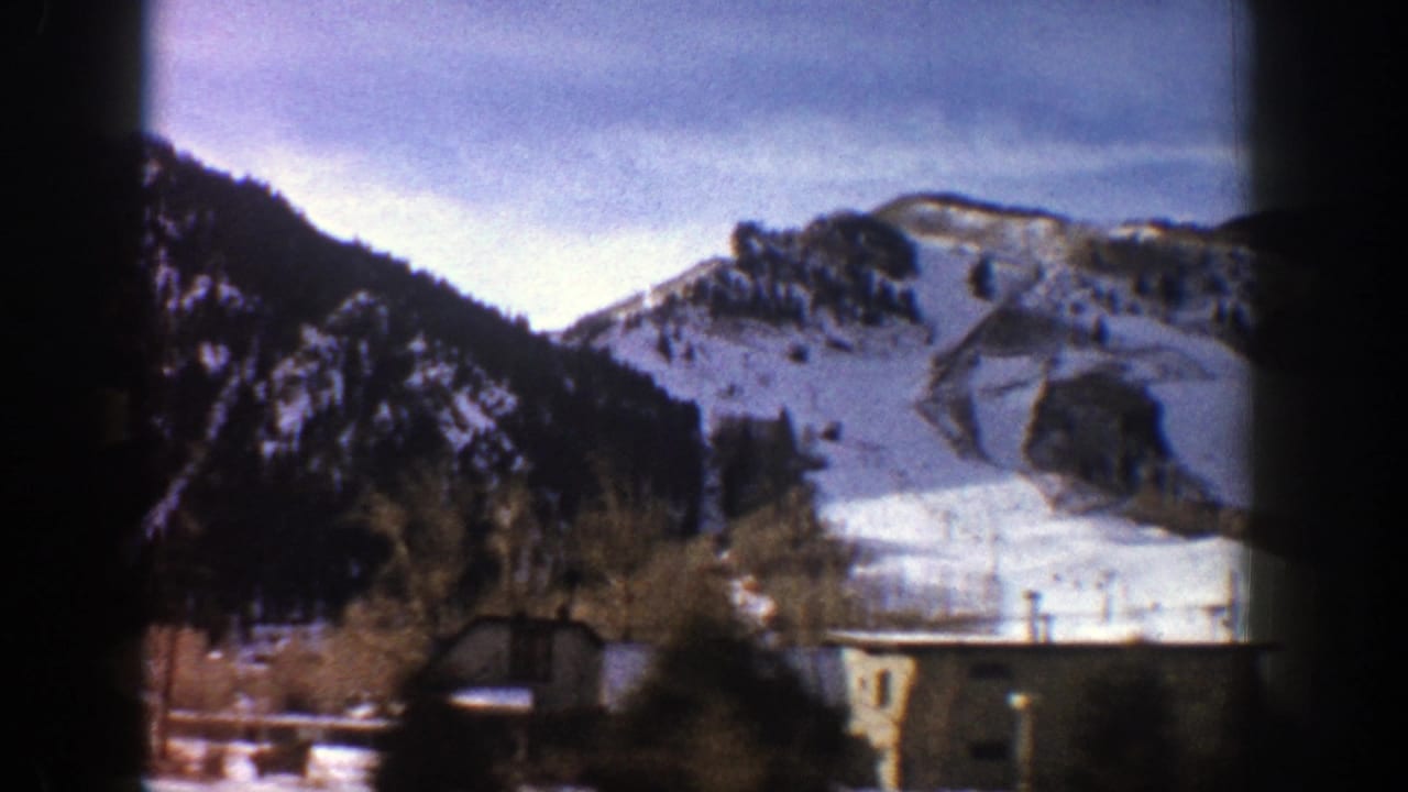 1961 Aspen, Colorado 8mm frame showing a snowy mountain slope, evergreen hillsides, foreground trees, and a small building.