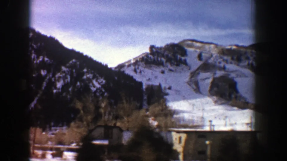 1961 Aspen, Colorado 8mm frame showing a snowy mountain slope, evergreen hillsides, foreground trees, and a small building.
