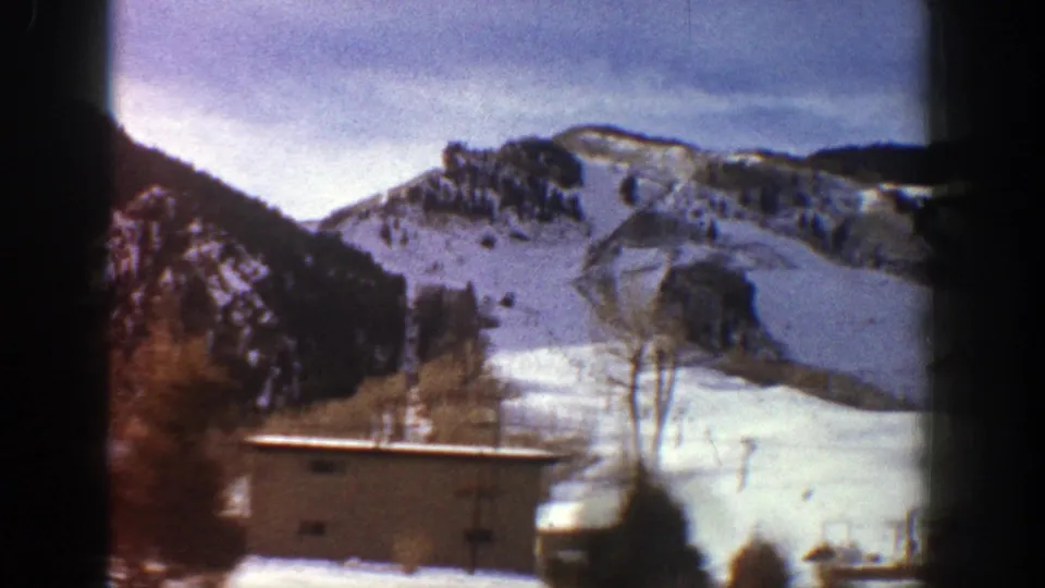 1961 Aspen, Colorado frame with snow-covered ski terrain, a rocky ridge, bare trees, and a low foreground building.