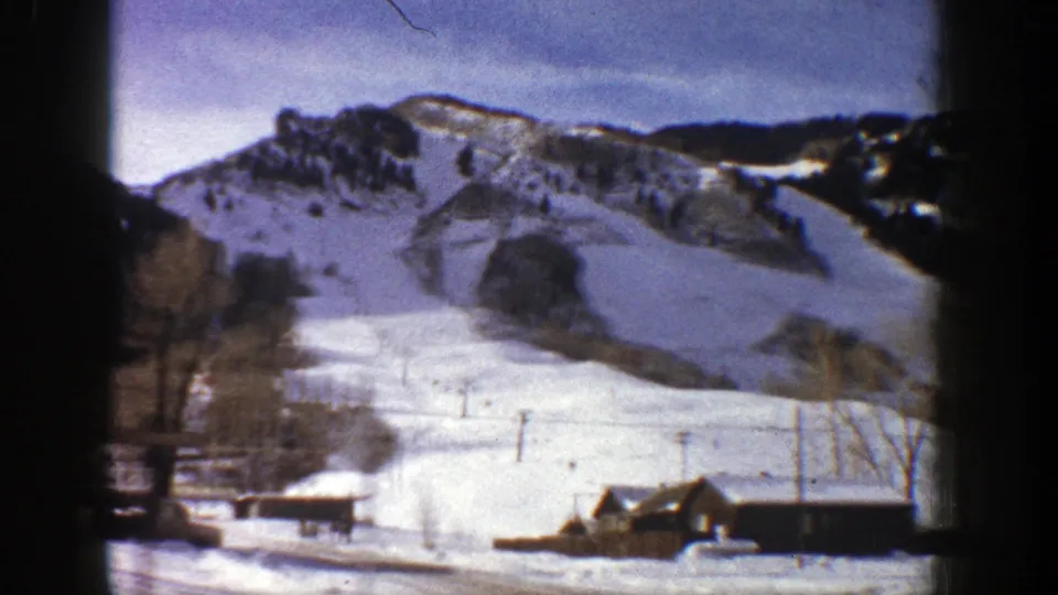 1961 Aspen, Colorado 8mm view of a snowy slope with utility poles, small cabins, bare trees, and dark wooded hills.