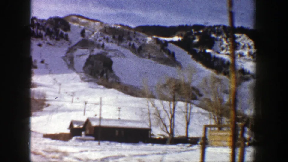 1961 Aspen, Colorado frame showing a snow-covered slope, small cabins, bare winter trees, utility poles, and an unreadable sign.
